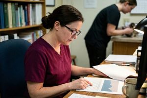 A veterinarian at her desk in lamplight reading a printed evaluation report on her practice, calculator at hand.